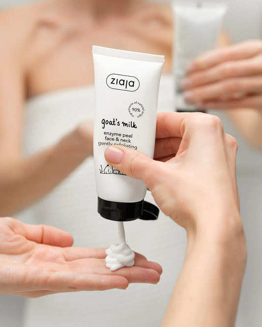 A hand holds a white tube of Ziaja Goal's Milk enzyme peel face & neck cream, squeezing a dollop of white cream into another hand, with a blurred bathroom mirror reflection in the background.
