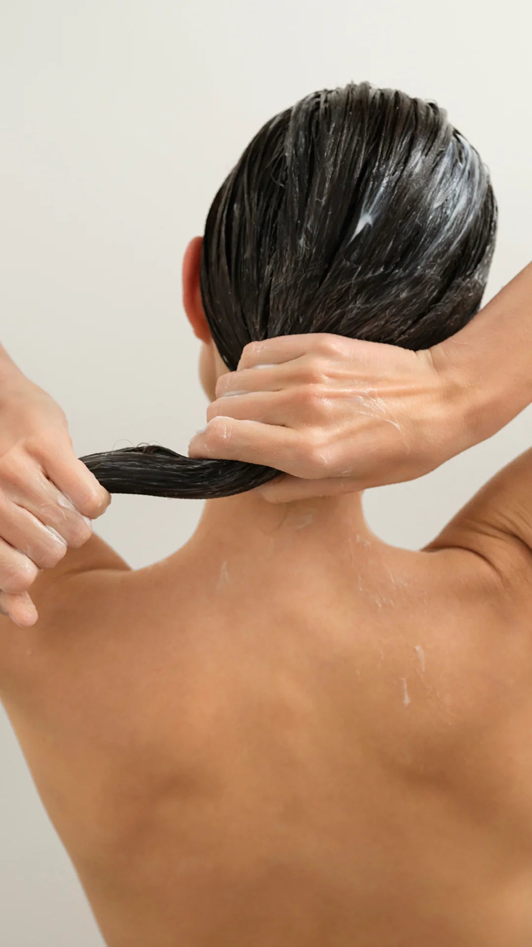 A close-up back view of a person with long, dark, wet hair being gently pulled down by hands, likely demonstrating a hair product or styling result against a plain light background.