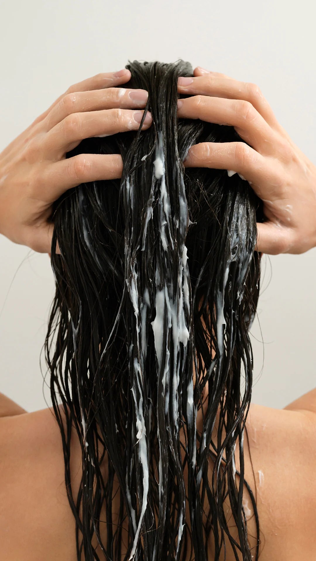 Back view of a person with long, wet black hair lathered with white shampoo, with hands massaging the scalp against a plain light background.