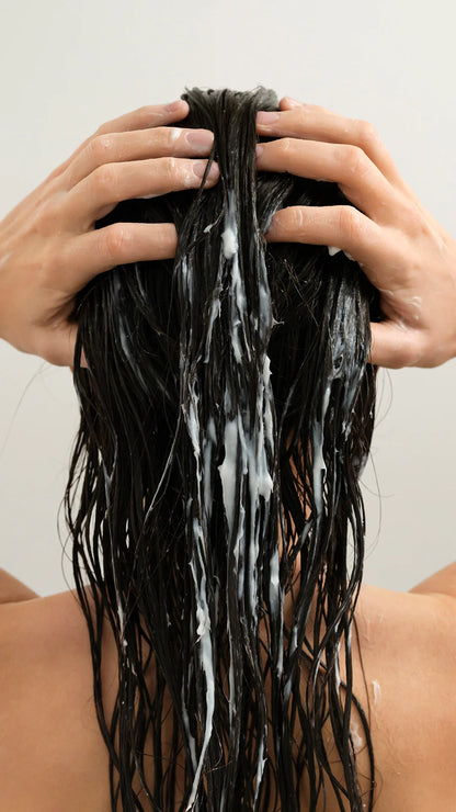 Back view of a person with long, wet black hair lathered with white shampoo, with hands massaging the scalp against a plain light background.